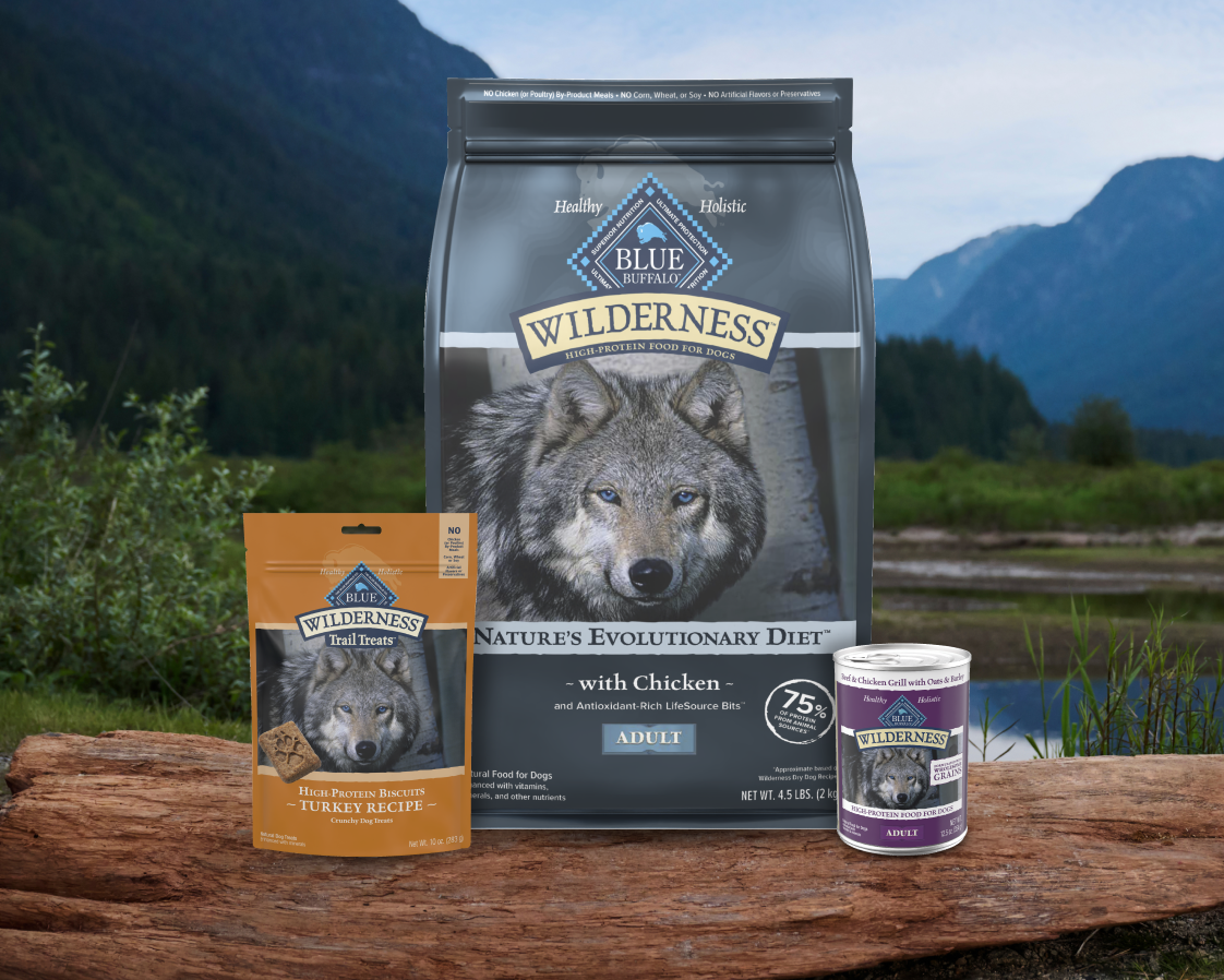 A bag of Wilderness dry dog food, a can of Wilderness wet dog food, and a pack of Wilderness dog treats are on a log outside.